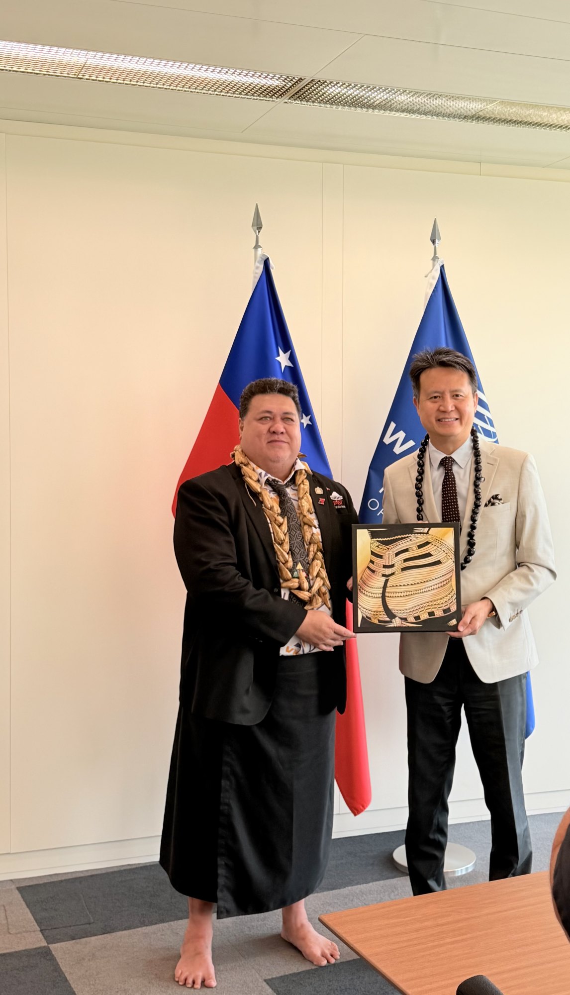 Samoa's Minister and WIPO Director General Daren Tang holding a framed painting of male tatau motifs presented as a cultural gift in front of the Samoa and WIPO flags